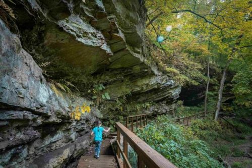 A person walks along a wooden path through a rocky, forested area with autumn foliage.