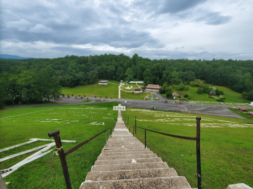 A view from the top of a staircase overlooking a green hillside and a parking area surrounded by trees under a cloudy sky.