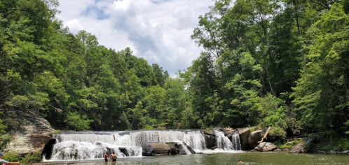 A serene waterfall surrounded by lush green trees, with people enjoying the water below under a partly cloudy sky.