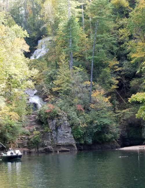 A serene lake surrounded by lush trees and a waterfall cascading down rocky cliffs. A small boat is visible on the water.