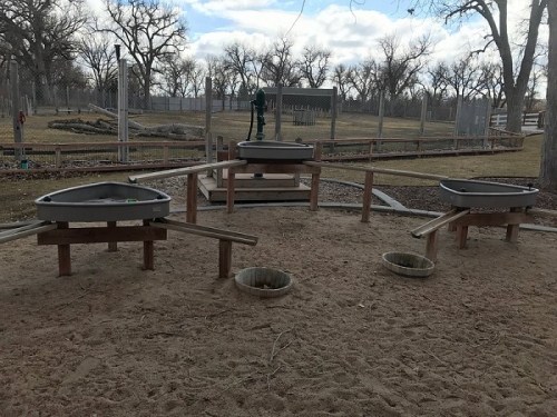 Three sand play areas with circular troughs and wooden supports in a park setting, surrounded by trees and open space.