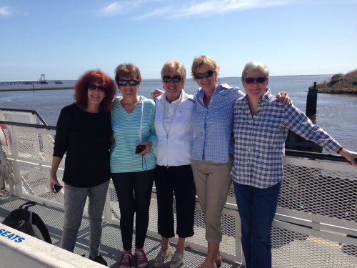 Five women stand together on a boat deck, smiling against a backdrop of water and a clear blue sky.