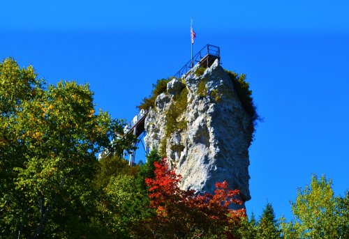 A tall rock formation topped with a flag, surrounded by trees and a clear blue sky.