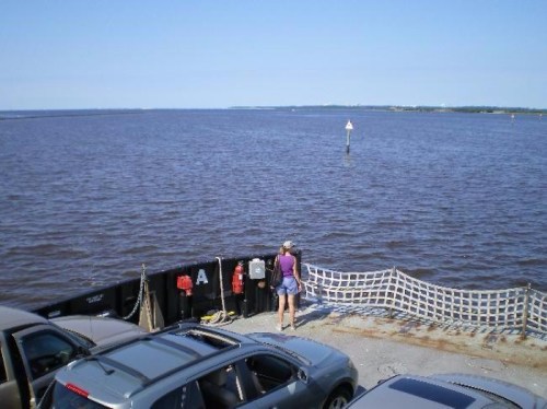 A person stands by the water's edge, overlooking a calm river with boats and a clear blue sky in the background.