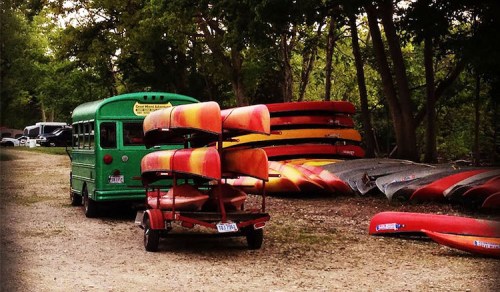 A green bus parked near stacked canoes and kayaks in a wooded area.