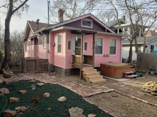 A pink house with a wooden porch, hot tub, and landscaped yard featuring stones and greenery.
