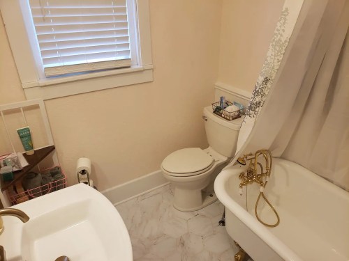 A bathroom featuring a toilet, a vintage bathtub, and a sink with a wooden shelf holding toiletries.