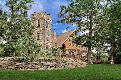 A stone tower and a wooden house surrounded by trees and greenery under a blue sky.