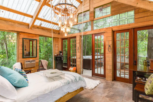 Cozy wooden bedroom with large windows, a chandelier, and a view of greenery, featuring a bed and seating area.