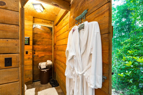 A cozy wooden bathroom with a white robe hanging, a mirror, and a rustic decor, surrounded by greenery.