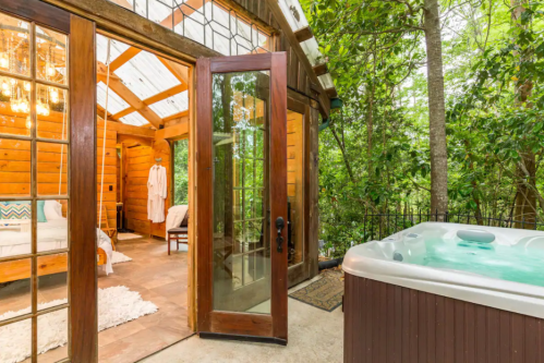 Cozy cabin interior with a hot tub outside, surrounded by lush greenery and large glass doors.