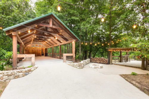 A rustic outdoor pavilion with string lights, surrounded by trees and a stone pathway.