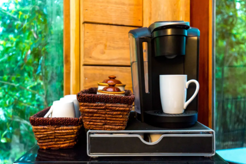 A coffee maker next to baskets of coffee pods and a white mug, set against a wooden backdrop with greenery outside.