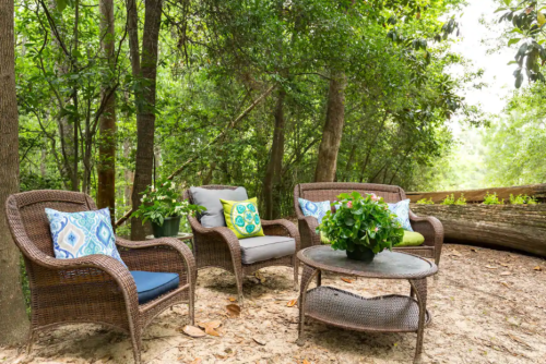 Cozy outdoor seating area with wicker chairs, colorful cushions, and a table, surrounded by lush greenery.