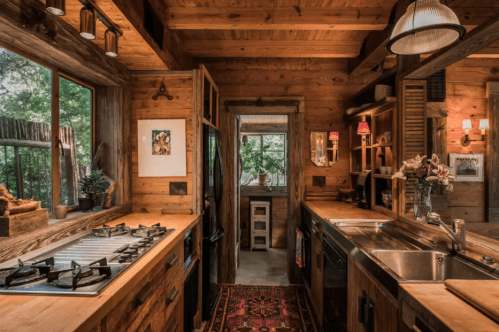 Cozy wooden kitchen with a gas stove, sink, and natural light from large windows, featuring rustic decor and plants.