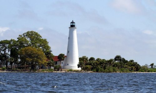 A white lighthouse stands on a small island, surrounded by water and greenery under a partly cloudy sky.