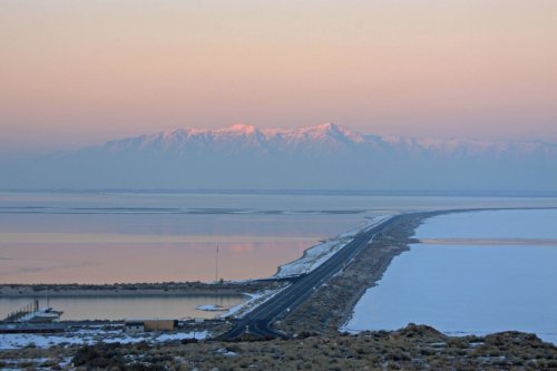 A serene landscape featuring a road leading to snow-capped mountains, with water reflecting soft pastel colors at sunrise.