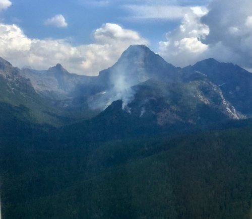 A mountainous landscape with smoke rising from a forested area under a cloudy sky.