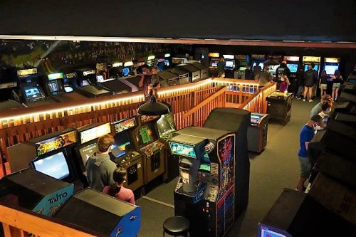 A retro arcade filled with vintage gaming machines and players enjoying various games in a dimly lit space.