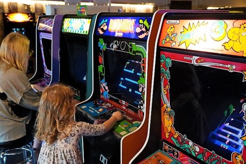 A child and an adult play retro arcade games in a colorful arcade setting.
