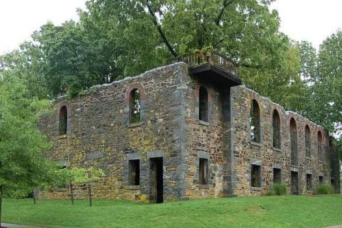 Ruins of a stone building surrounded by greenery, featuring arched windows and a balcony on the upper level.