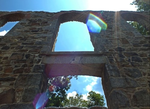 Sunlight streams through the arched windows of a stone ruin, highlighting the textured walls and surrounding greenery.