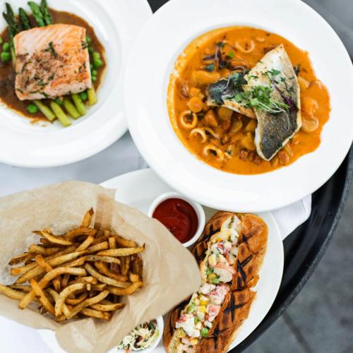 A table with a plate of salmon, seafood pasta, a hot dog with toppings, and a basket of fries with ketchup.