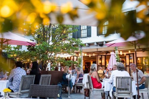 Outdoor dining scene with people at tables, surrounded by greenery and warm lighting in a cozy restaurant setting.