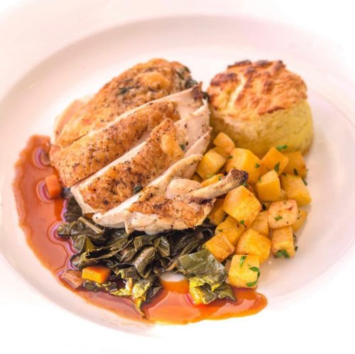 A plate of sliced roasted chicken with collard greens, roasted squash, and a cornbread muffin, drizzled with sauce.