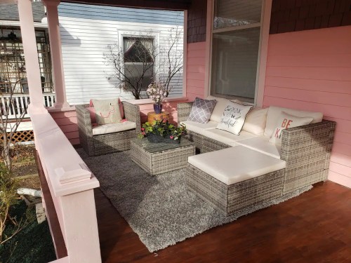 Cozy porch with wicker furniture, cushions, a coffee table, and decorative plants on a soft rug against a pink wall.