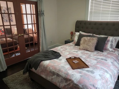 Cozy bedroom with a patterned bedspread, a tray on the bed, and a view of a living room through glass doors.