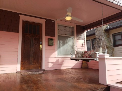 A cozy porch with a wooden swing, floral cushions, and a pink exterior, featuring a brown door and wooden flooring.