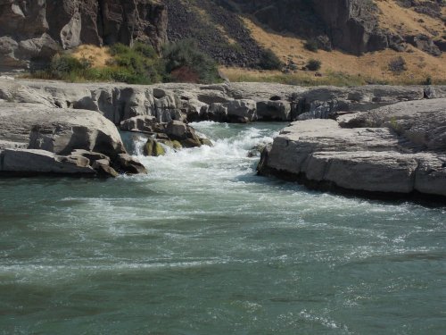 A flowing river surrounded by rocky terrain and sparse vegetation under a clear sky.