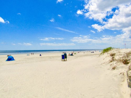 A sunny beach scene with people relaxing on the sand, blue sky, and gentle waves in the background.