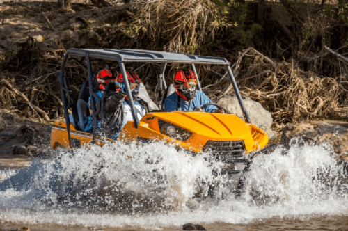 A bright orange off-road vehicle splashes through water, carrying four people wearing helmets in a rugged outdoor setting.