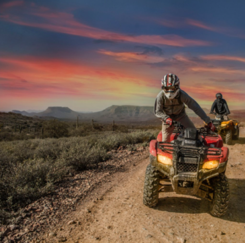 Two riders on ATVs navigate a dirt path against a vibrant sunset in a desert landscape.