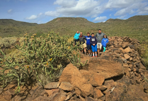 A group of six people stands on rocky terrain with cacti and mountains in the background under a partly cloudy sky.