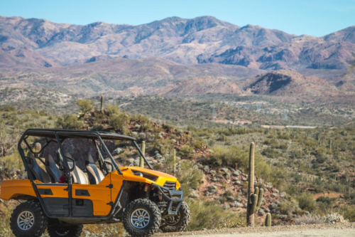 An orange off-road vehicle parked on a dirt path with mountains and cacti in the background.