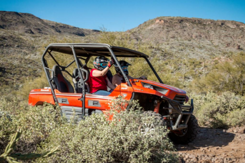 Two people in an orange off-road vehicle navigate a rocky trail in a desert landscape.