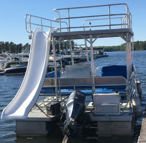 A pontoon boat with a slide, docked by the water, surrounded by other boats in the background.