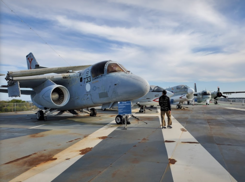 A person stands near military aircraft on a flight deck, with several planes visible under a blue sky.