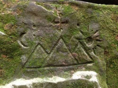 Ancient rock carving featuring triangular mountains, a sun, and celestial symbols, covered in moss.