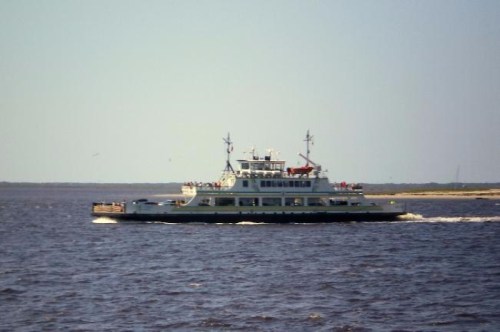 A ferry boat sails across a calm body of water on a clear day, with a shoreline visible in the background.