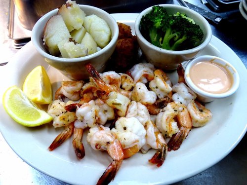 A plate of shrimp with lemon wedges, baked potatoes, broccoli, and a dipping sauce on the side.