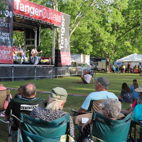 A crowd enjoys a live band performance on stage at an outdoor event, surrounded by trees and vendor tents.