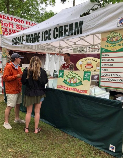 A food stall with a sign for homemade ice cream, featuring two customers ordering from a vendor under a tent.
