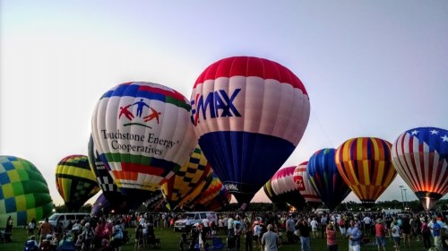 Colorful hot air balloons fill the sky at a festival, with a crowd gathered below in a grassy field at dusk.