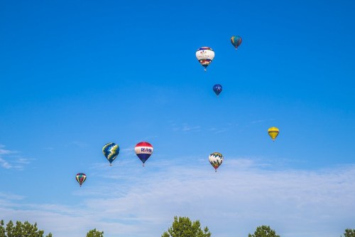 A clear blue sky filled with colorful hot air balloons floating above green trees.