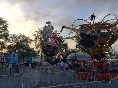A carnival scene at sunset featuring rides, including a Ferris wheel and a spinning attraction, with people enjoying the festivities.
