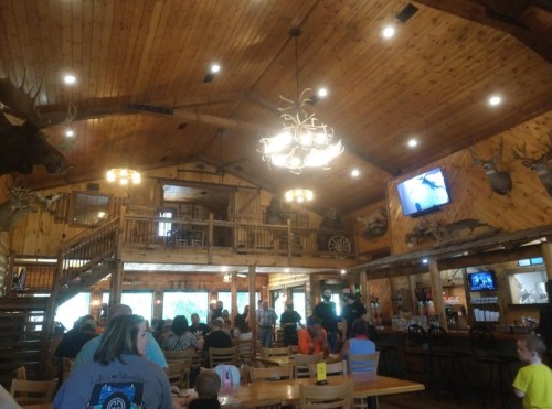 A rustic restaurant interior with wooden decor, antler chandeliers, and patrons seated at tables.